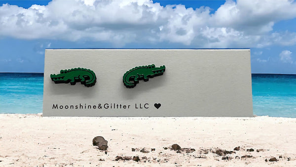 May include: Two green alligator earrings on a white card with the text &#39;Moonshine&Glitter LLC&#39; and a heart symbol.